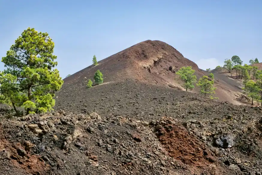 Cono volcánico de laderas oscuras y pardas con pinos dispersos y piedra volcánica rojiza en primer plano, bajo un cielo azul despejado