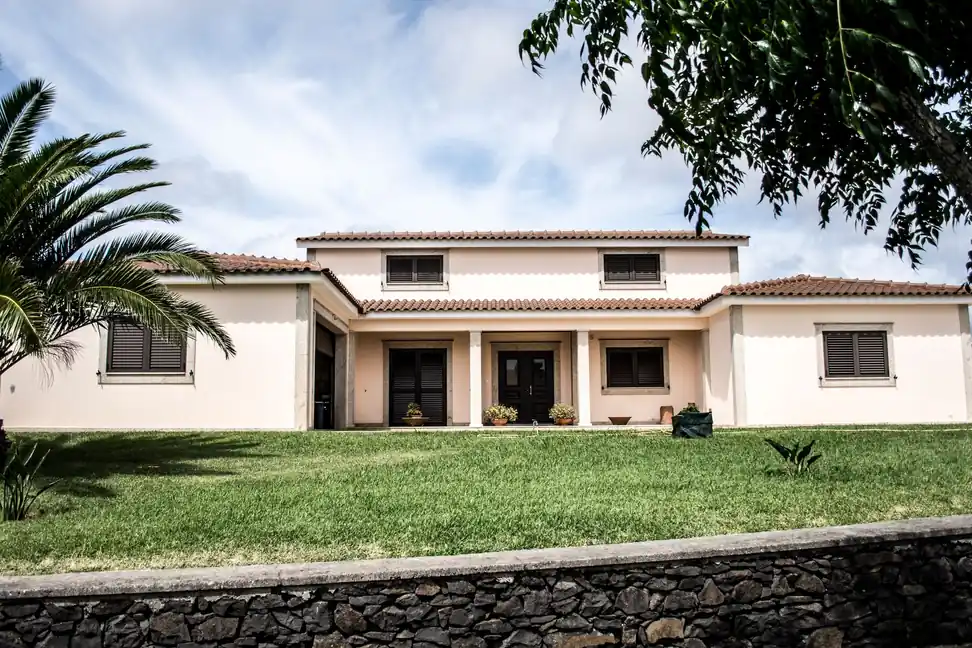 Casa unifamiliar de dos plantas con tejado de teja, jardín de césped y palmera tras un muro de piedra
