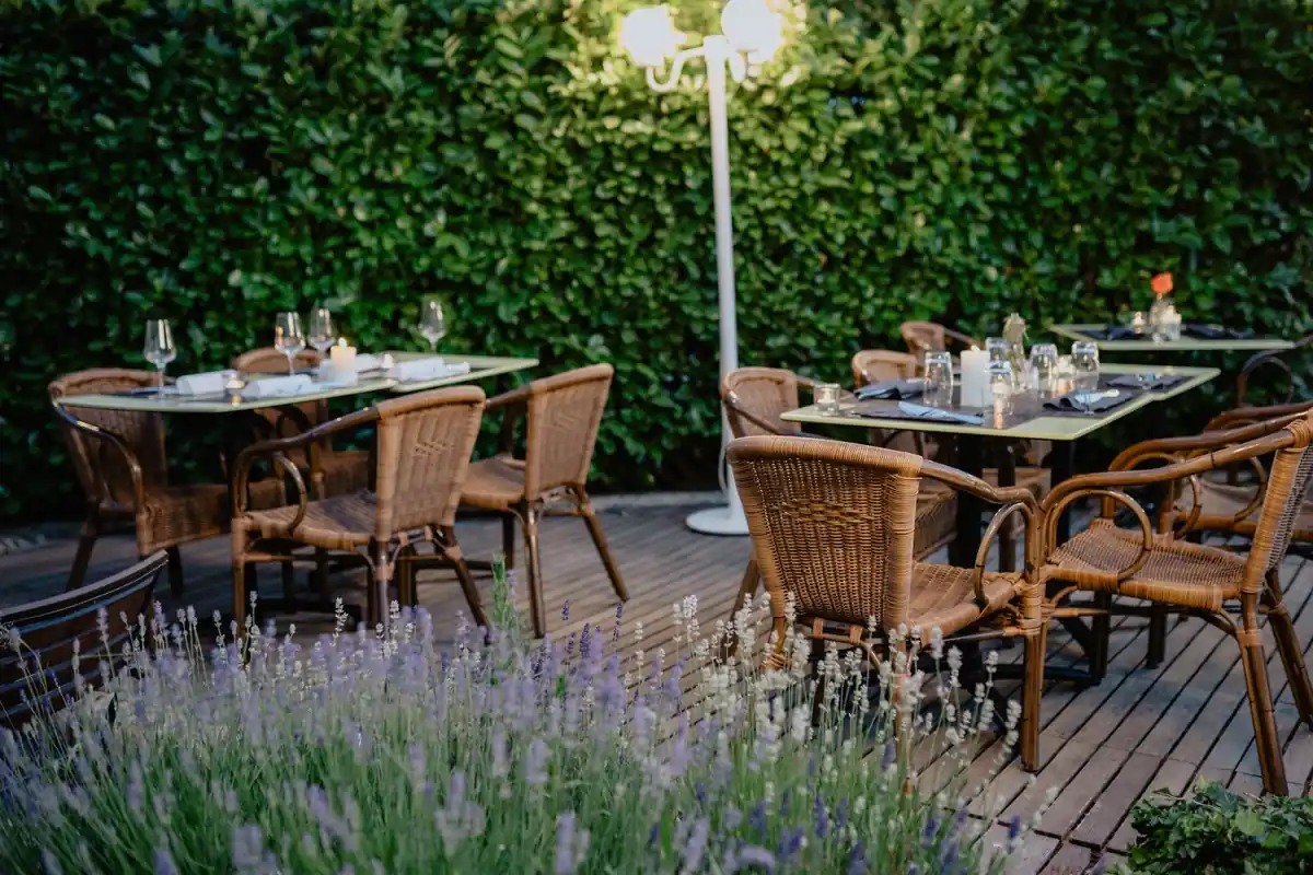 Terraza de un restaurante con las mesas puestas, sillas de mimbre vacías y una farola junto a un seto