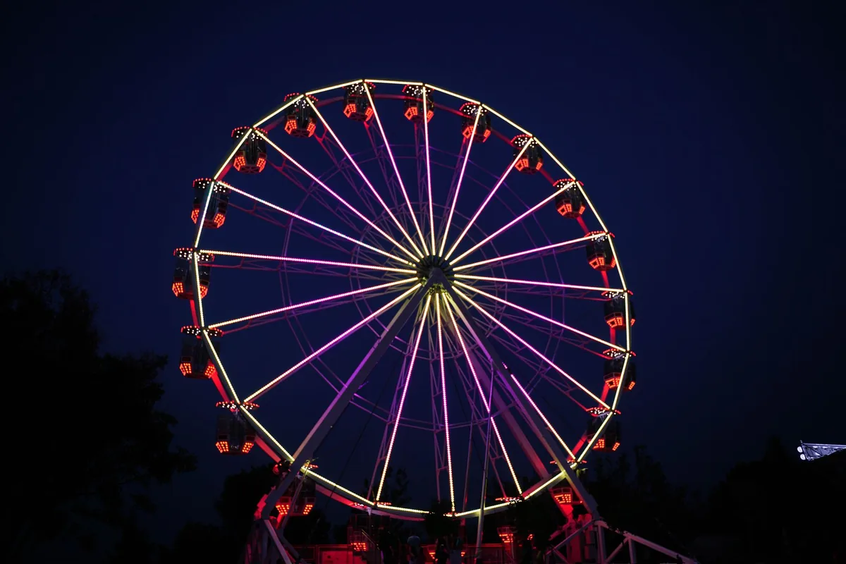 Noria de feria iluminada de noche, con radios de luz morada y amarilla y cabinas con luces rojas