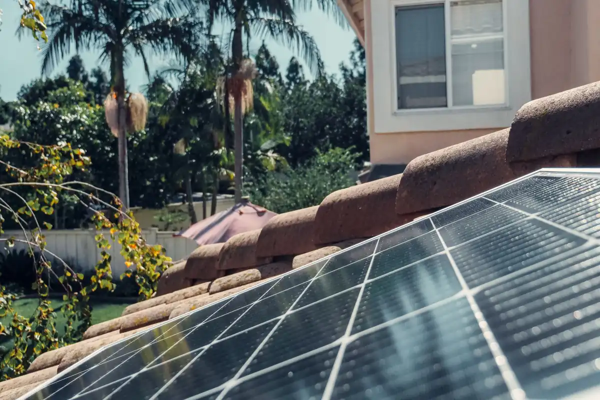 Placa solar instalada sobre un tejado de teja curva, con palmeras al fondo y una casa de fachada rosada