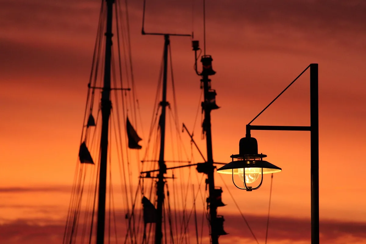 Farola encendida junto a los mástiles de unos barcos, con el cielo anaranjado del atardecer