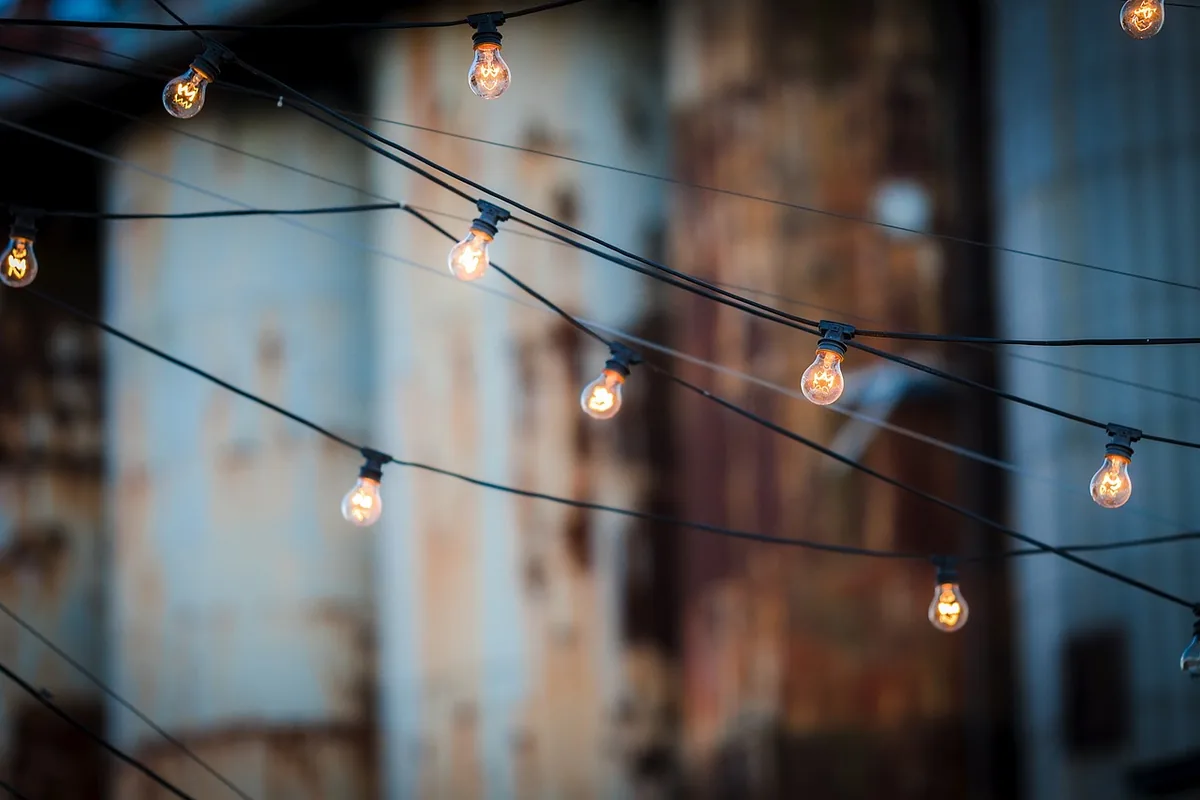 Guirnalda de bombillas encendidas colgada de cables al aire libre, con fachadas desenfocadas al fondo